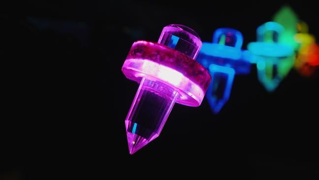A close-up shot of a clear, double-terminated quartz crystal used for crystal healing, glowing with vibrant purple light, held by a circular ring casing against a black background with other blurred, colorful lights.