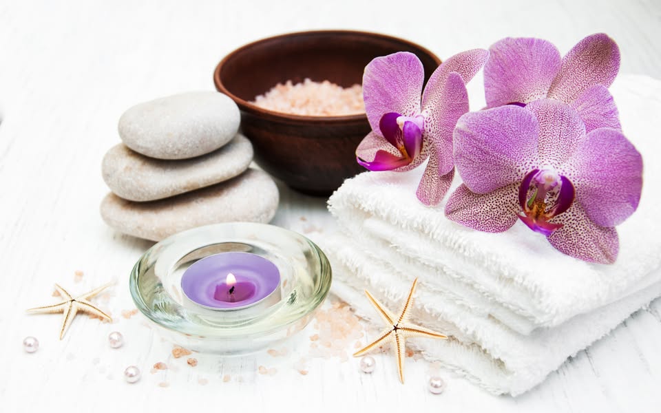 Wellness arrangement featuring a cluster of purple orchid blossoms resting on stacked white spa towels with massage stones and a lit purple candle.